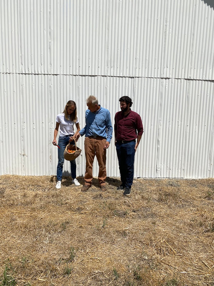 Three people standing in front of a corrugated metal wall on a dry, grassy area.