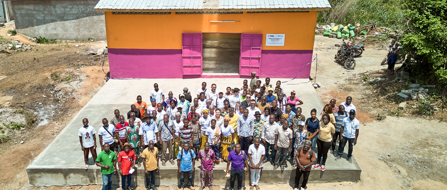 Group of people standing in front of a colorful building with a dirt road and greenery in the background.