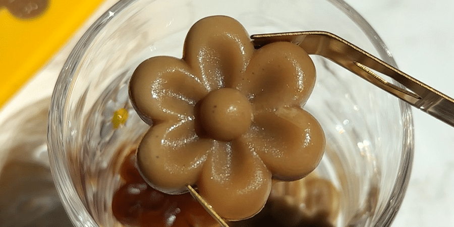 Golden tongs holding a flower shaped coffee ice cube over a glass.